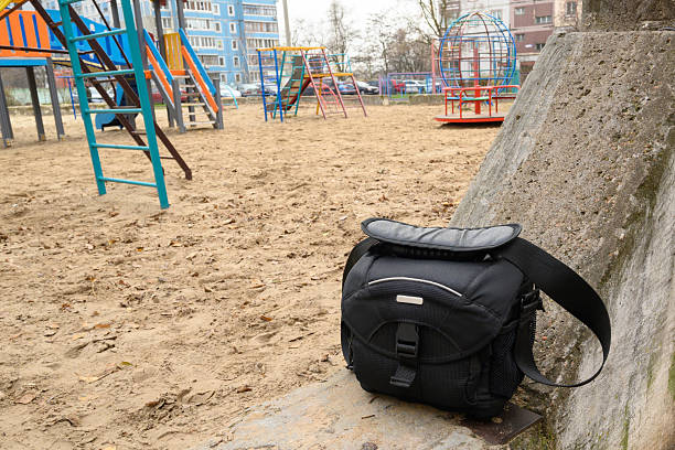 a black textile bag for a camera or camcorder stands on a concrete block at the edge of a playground, with sand and play structures in the background