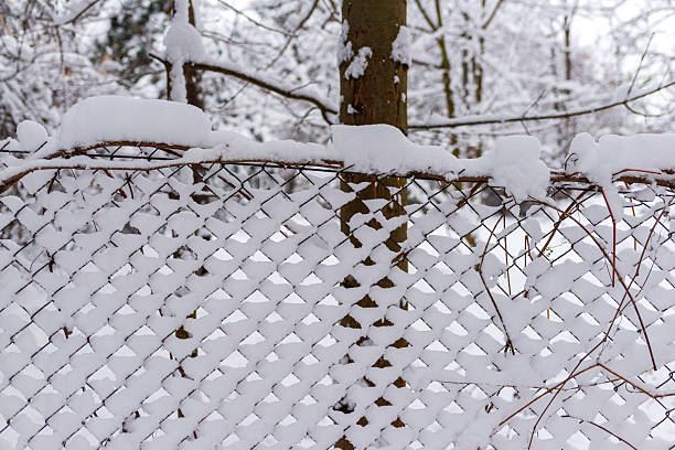 A metal chain-link fence is densely packed with fresh snow, creating a geometric winter texture. Soft white layers contrast with dark branches and tree trunks, forming a calm seasonal scene perfect for backgrounds and festive visuals.