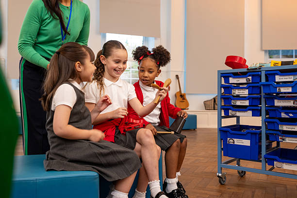 Medium shot of an unrecognisable music teacher with a small group of young schoolchildren in a school hall in Northumberland, North East England. They are playing with different musical instruments, smiling and talking while the teacher assists the students.Videos similar to this scenario available.