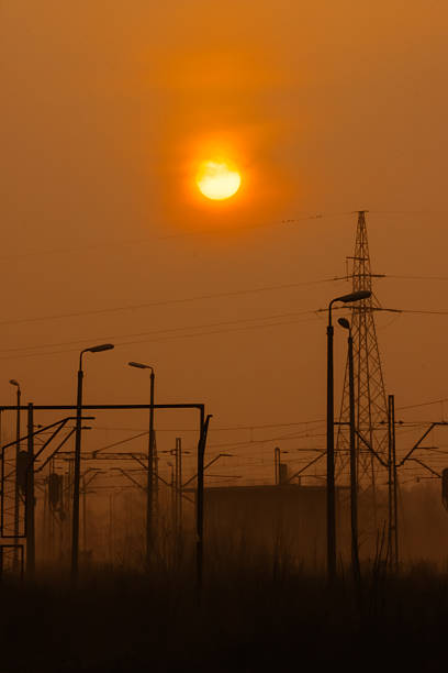 A dramatic scene of railway infrastructure silhouetted against an intense orange sunset. Power lines, signal poles and metal structures fade into a hazy industrial landscape illuminated by warm evening light. The atmospheric conditions create a moody, cinematic feel, highlighting the contrast between nature and urban technology.