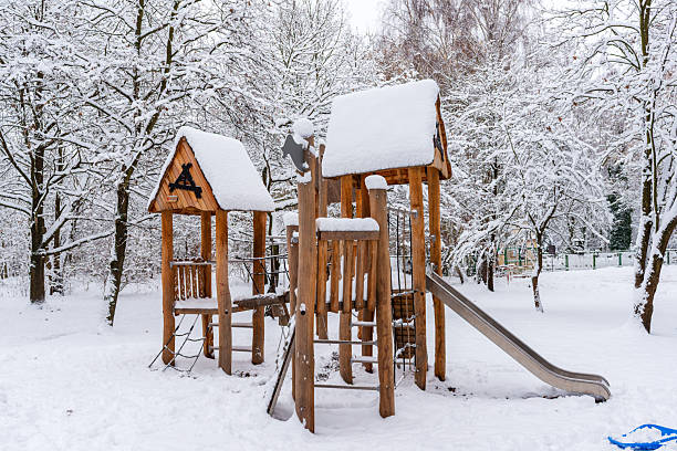Natural wooden play towers stand quiet under fresh snow, surrounded by frosted trees. The blend of warm wood and cold winter tones creates a cozy seasonal atmosphere reminiscent of holiday postcards.