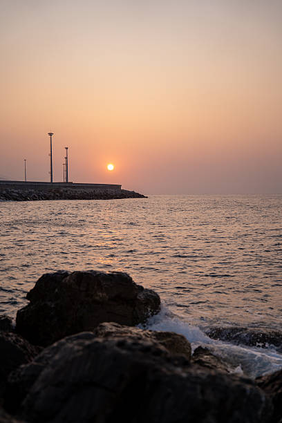 Sunset over rocky shore with foreground boulders and calm sea, glowing orange sky and gentle surf washing pebbled coastline,