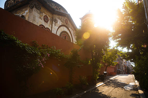 A serene Istanbul street bathed in warm golden-hour light. The sun shines softly behind the dome of a historic church, casting gentle flares across the leafy trees and terracotta wall. Vibrant plants line the walkway, adding natural beauty to the peaceful residential setting. This atmospheric image blends architectural charm, soft sunlight, and a calm neighborhood mood.