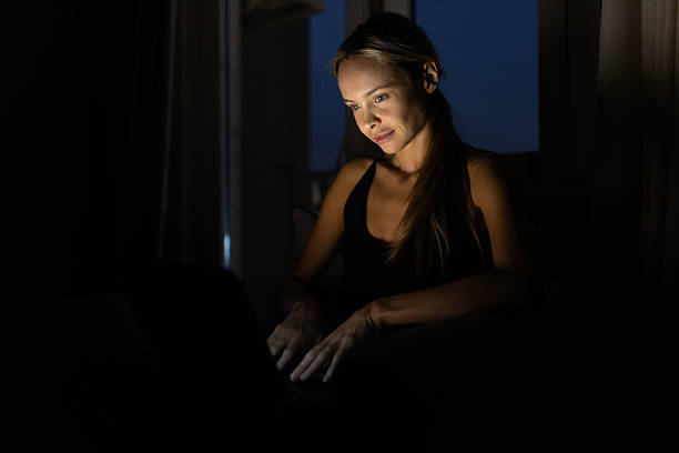Beautiful woman, freelancer, working from home at night. Sitting on the armchair in her living room and working on laptop in dark room