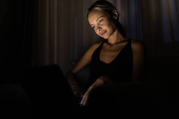 Beautiful woman, freelancer, working from home at night. Sitting on the armchair in her living room and working on laptop in dark room
