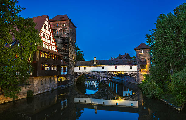 Twilight scene featuring a historic stone bridge and timber-framed buildings in Nuremberg, Bavaria. The calm canal reflects the architecture, creating a serene, picturesque view of old-world German charm.