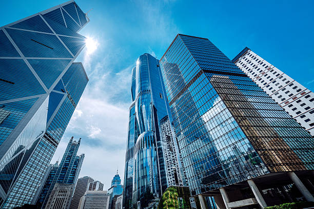 A dramatic view of Hong Kong's glass-clad skyscrapers reaching toward a bright blue sky. The dynamic angle emphasizes urban growth, modern architecture, and vibrant city life.