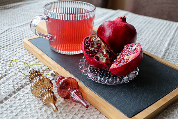 Close-up of raspberry red tea in a glass mug with fresh pomegranate slices placed on a slate tray. Decorative Christmas ornaments add festive mood. Soft textured fabric background.