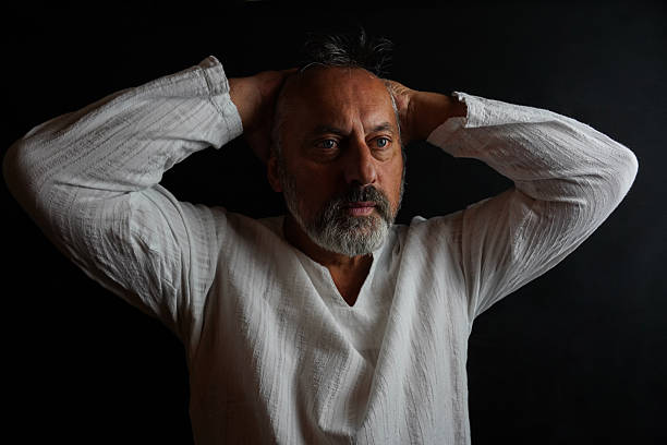 A middle-aged man poses in a studio setting against a dark black background. He is wearing a white linen shirt while taking various poses – looking to the side, crossing his arms, thinking, and holding his hands together in contemplative positions. The images represent portrait poses, introspection, and a calm expression under controlled studio lighting.