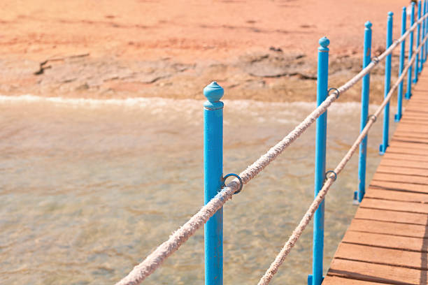 Wooden walkway features blue railing and rope, leading to calm water, evoking a peaceful ambiance for relaxation and leisure by the beach during sunset