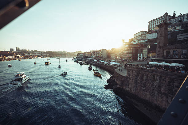 Panoramic aerial view of the Douro River in Porto, Portugal, with traditional Rabelo boats moored along the waterfront and the historic Ribeira district on the right. The sun glistens on the water, creating a scenic urban landscape popular with tourists.