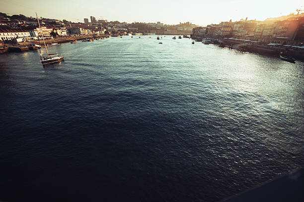 Panoramic aerial view of the Douro River in Porto, Portugal, with traditional Rabelo boats moored along the waterfront and the historic Ribeira district on the right. The sun glistens on the water, creating a scenic urban landscape popular with tourists.