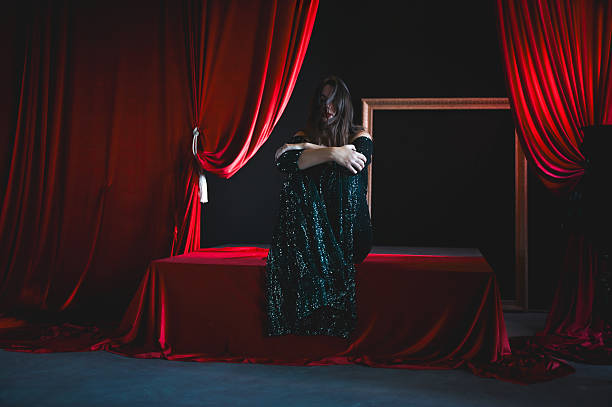 Woman sitting on a stage with red curtains, hugging knees, experiencing sadness or loneliness