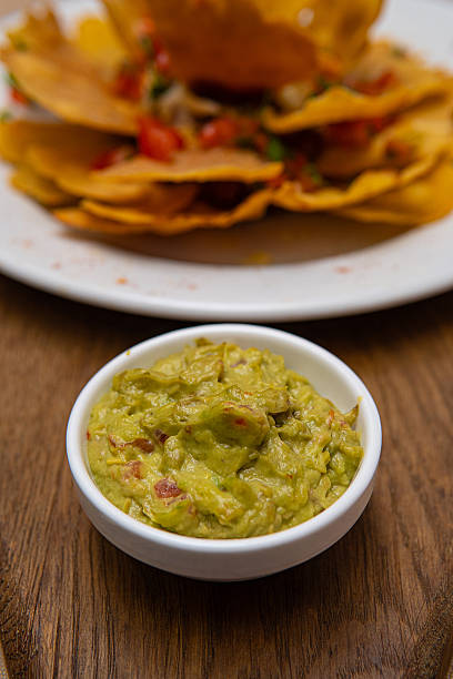 Fresh guacamole dip in a white bowl on a wooden table with blurred tortilla chips behind.