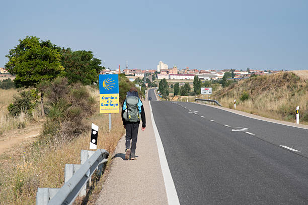 Sahagun is located at the midpoint of the French leg of the Camino de Santiago pilgrimage. A male pilgrim walks along an asphalt road toward Sahagun, visible in the distance. Along the road stands a sign pointing to the Camino de Santiago pilgrimage route.