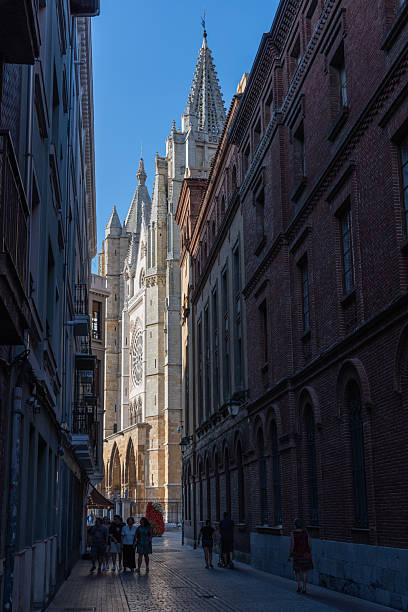 At the end of an alleyway between the buildings of downtown Leon, the spire of the Leon Cathedral and the entrance to the Leon Cathedral are seen obliquely, pouring into the blue sky.