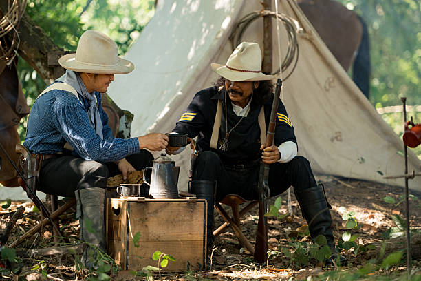 Two cowboys sit at a rustic campsite, sharing a cup of coffee beside a wooden crate and brewing pot