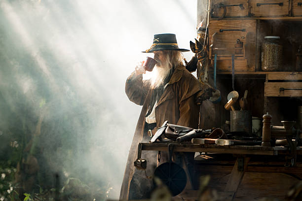 A senior cowboy stands beside a food cart, drinking coffee while morning light beams through the smoky forest