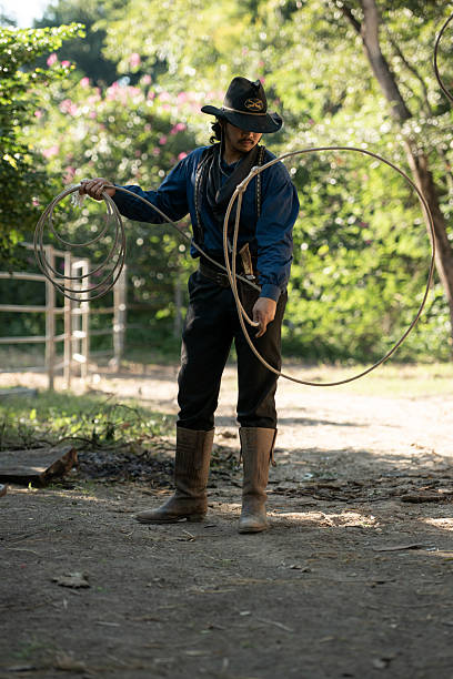 A cowboy handling a lasso rope for training at a rustic camp setup with wooden storage and cooking tools in the background