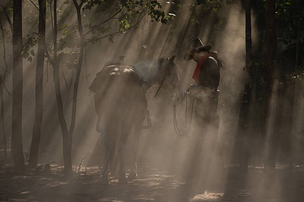 A cowboy leading his saddled horse through a dusty forest as sun rays stream through the trees