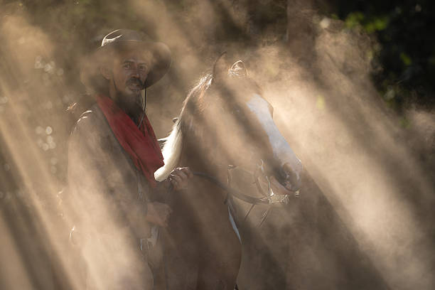 A cowboy leading his saddled horse through a dusty forest as sun rays stream through the trees