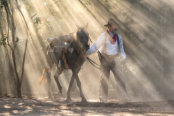 A cowboy leading his saddled horse through a dusty forest as sun rays stream through the trees