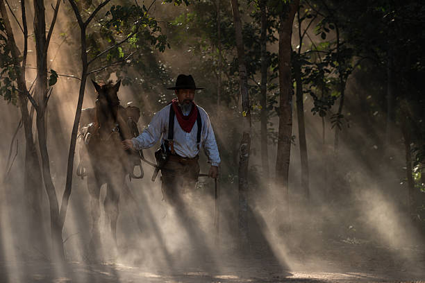 A cowboy leading his saddled horse through a dusty forest as sun rays stream through the trees