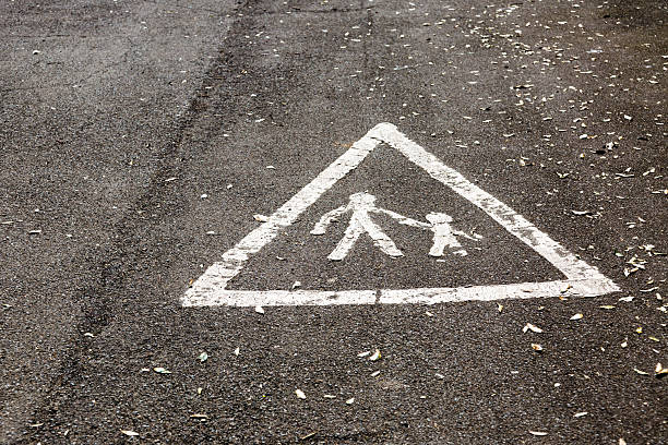 A white pedestrian crossing marking inside a triangular warning sign painted on wet asphalt. The scene shows two figures holding hands, emphasizing street safety for children and families.