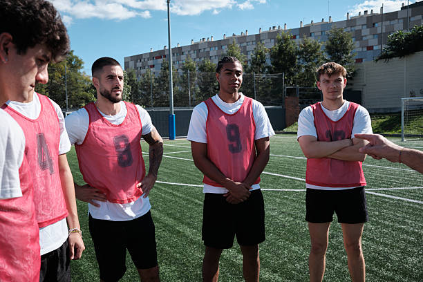 Young adult soccer players on a green turf field listening intently to their coach, receiving instructions during training