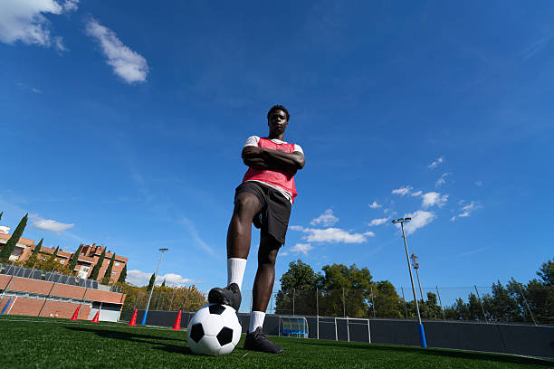 One young adult male football player, arms crossed, standing confidently with a foot on a soccer ball on a green pitch