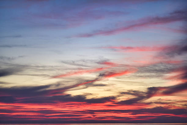 Red and pink streaks of sunset clouds spread across a soft pastel sky above the calm sea. Gentle color transitions create a peaceful evening atmosphere.