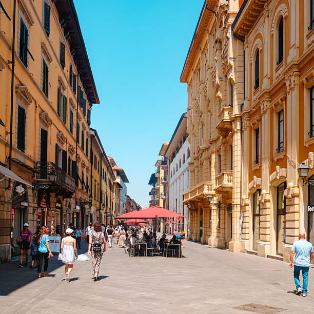 Pisa, Italy - June 26, 2025: A lively view of Corso Italia shows shoppers and pedestrians moving between the warm toned facades and the outdoor cafe tables, shaded by bright red umbrellas