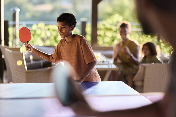 Happy African American boy playing table tennis against someone while mother and girl are in the background.