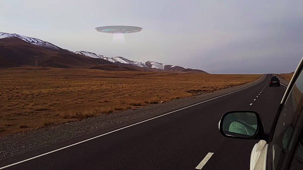 An unidentified flying object is seen hovering over a remote highway landscape surrounded by wide plains and distant peaks.