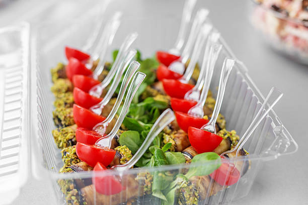 Close-up of pistachio-coated eggplant rolls with cherry tomato halves, served in plastic forks over greens, ready for catering delivery.