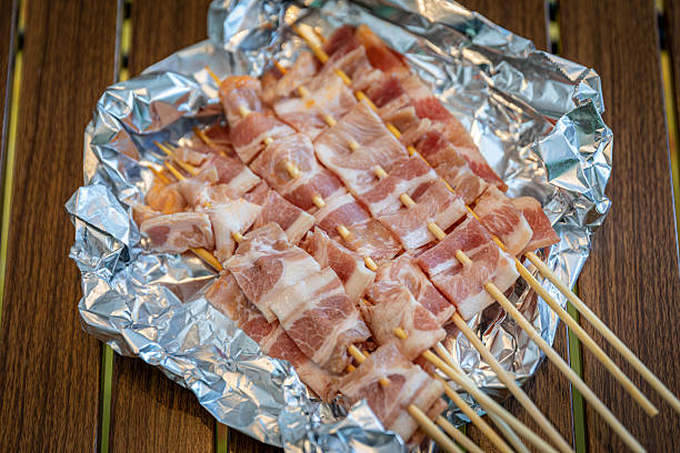 A folding wooden table on grass (or artificial turf) is loaded with picnic and barbecue foods — pizza in a box, skewers of meat and seafood, bread, a bowl of cooked crawfish, fruit, and a bottle of juice. A cooler, portable grill and two folding chairs sit nearby, creating a relaxed gathering or camping-party scene