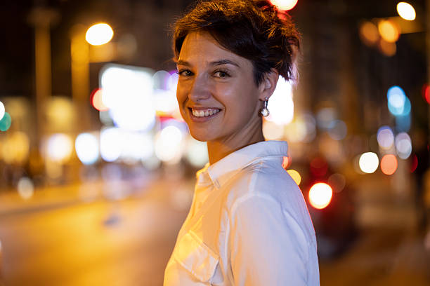 A young woman stands confidently in a brightly lit city street at night, captured with a shallow depth of field that creates colorful bokeh lights in the background. She smiles naturally while wearing a white shirt, giving the scene a warm and expressive urban atmosphere.