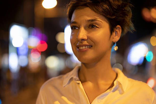 A young woman stands confidently in a brightly lit city street at night, captured with a shallow depth of field that creates colorful bokeh lights in the background. She smiles naturally while wearing a white shirt, giving the scene a warm and expressive urban atmosphere.