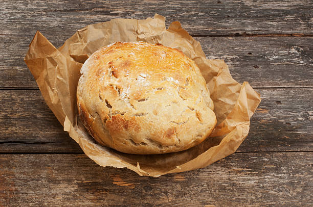 Single homemade sourdough bread wrapped in baking paper resting on a dark wood bench