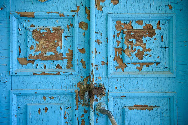 Close-up of a weathered wooden door with faded blue paint flaking off in large patches. The symmetrical panel design and rusted metal latch evoke tactile decay, quiet abandonment, and the layered passage of time.