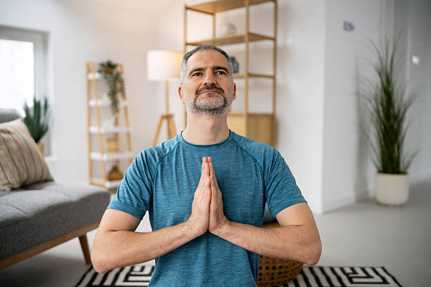 A middle-aged man is practicing yoga in a modern living space, with hands in a prayer position. The room features a stylish bookshelf and a cozy atmosphere, suggesting relaxation and mindfulness.
