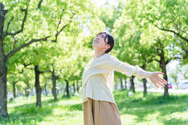 A woman stands in a sunlit park surrounded by fresh green trees, spreading her arms wide as she takes a deep, refreshing breath.The soft sunlight and vibrant greenery create a serene image that conveys themes of relaxation, wellness, mindfulness, and connection with nature.