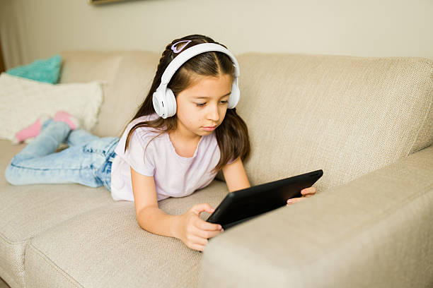 istock Little girl wearing headphones and using a digital tablet for e-learning or entertainment while relaxing on a sofa at home 2248876974