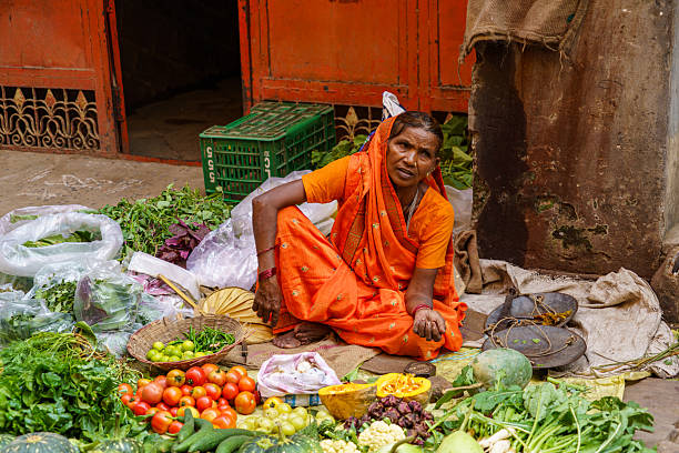 India. Uttar Pradesh state. Varanasi (Benares). A vegetable vendor sits behind her makeshift stall, selling fresh fruits and vegetables on a street in Varanasi (Benares). The woman wears a bright orange sari, a color often associated with spirituality and tradition in India