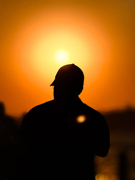 Istanbul, Turkey, 2 September 2025, A dramatic portrait featuring the silhouette of a person wearing a cap, positioned against the bright, circular glow of a setting sun. The warm orange and golden hues of the sky create a powerful, contemplative atmosphere, symbolizing reflection, the end of a day, or a peaceful moment in nature.