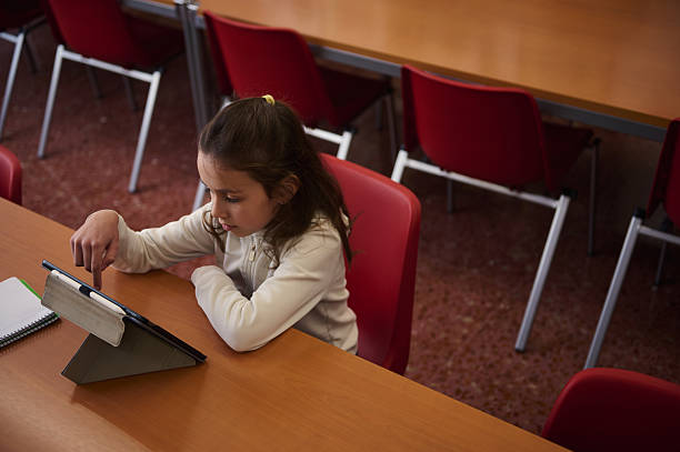 A young girl sits at a classroom desk, focused on a tablet in a school library, conveying learning, technology use, and study.