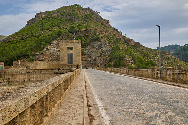 Scenic road built along the top of a massive stone dam, surrounded by mountain hills and greenery in Spain.