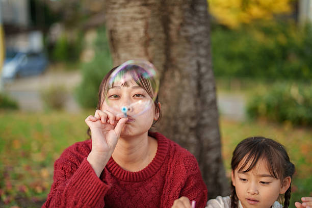 Asian mother blowing a soap bubble while sitting outdoors with her young daughter beside her in an autumn park. Natural family moment showing calm playtime, gentle presence, and everyday outdoor lifestyle with soft natural light and seasonal colors.