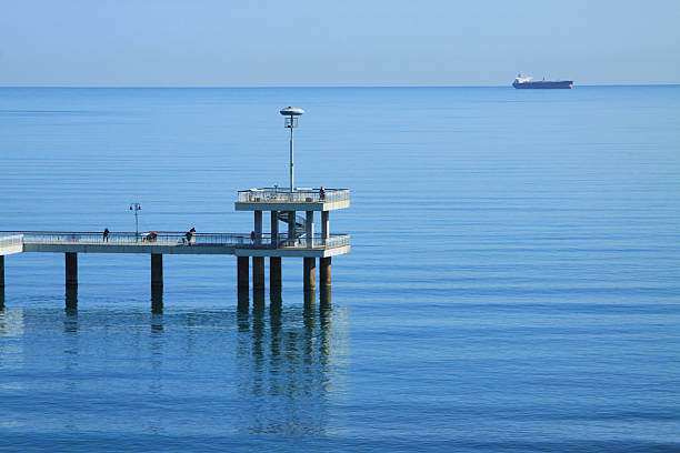 Seaside pier stretches over tranquil blue waters, with gentle waves and a distant ship, evoking a peaceful coastal environment for leisure