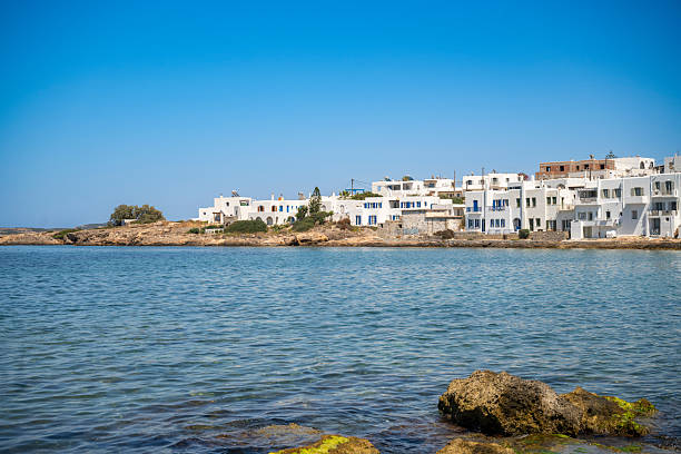 Whitewashed buildings with blue accents line the calm waterfront in Naousa on Paros island, Greece, captured on a bright sunny day with clear waters in the foreground.